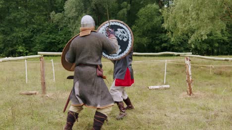 Two-medieval-warriors-in-armor-fighting-with-shield-and-sword-on-a-green-field-in-slow-motion