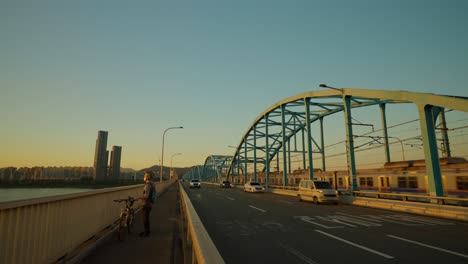 Dongjak-bridge-traffic-and-metro-line-4-train-passing-at-sunset,-Unrecognizable-rider-with-bicycle-stopped-to-enjoy-the-sun-setting-view-from-bridge-walkway-,-Seoul,-South-korea