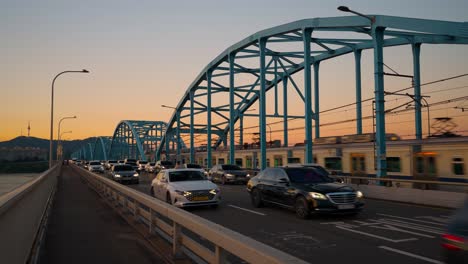 Two-Seoul-metro-trains-are-going-in-opposite-directions-on-Dongjak-bridge-at-sunset,-and-many-cars-are-in-traffic-on-the-bridge-road,-South-Korea