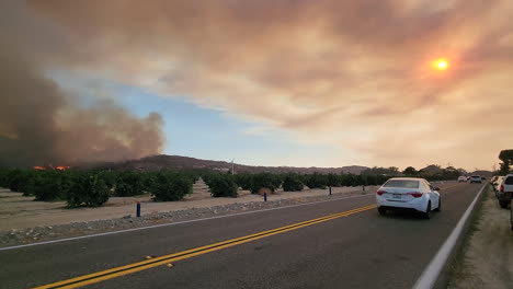 Car-traffic-next-to-the-Fairview-Fire,-burning-hill-and-thick-black-smoke-in-Hemet,-California