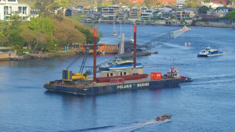 A-large-barge-carrying-a-crane-sails-up-the-river-flanked-by-river-city-catamarans