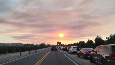 POV-shot-of-a-motor-vehicle-driving-along-a-congested-road,-the-shoulder-of-the-road-lined-with-parked-vehicles-as-people-view-the-devastation-caused-by-the-Fairview-fire-at-sunset,-Hemet,-California