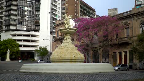 Slow-motion,-handheld-wide-view-of-Utopias-Fountain-in-Rosario,-Argentina,-against-a-beautiful-backdrop-of-buildings-and-a-blossomed-jacaranda-tree