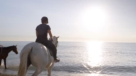 Rear-view-of-equestrian-with-helmet-riding-horses-into-ocean-water-during-sunset-light---Enjoy-good-feeling-underwater-in-summer