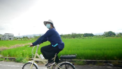 Asiatische-Frau-In-Blauem-Langschlafoutfit-Und-Mit-Gesichtsmaske,-Die-An-üppigen-Grünen-Reisfeldern-Vorbeireitet