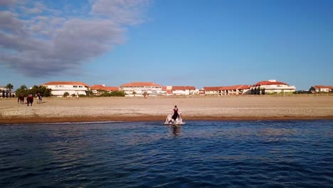 Drone-shot-of-female-equestrian-riding-horse-at-shore-of-beach-in-France---Hotel-buildings-in-background-In-St-cyprien