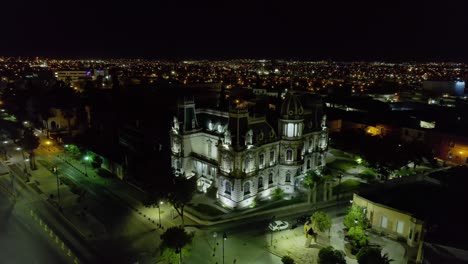 Illuminated-Quinta-Gameros-building-in-Chihuahua,-dark-evening-in-Mexico---approaching,-aerial-view