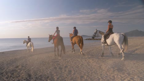 Group-of-young-women-riding-horses-at-beach-towards-ocean-and-enjoying-sunset-golden-hour