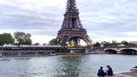 Couple-Hang-Out-On-The-Bank-Of-Seine-River-Near-The-Pont-d'Iéna-And-Eiffel-Tower-In-Paris,-France