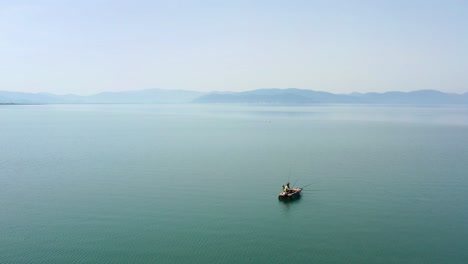 Totaler-Zen-Moment-Für-Einen-Fischer-In-Einsamkeit-In-Seinem-Kleinen-Fischerboot-Inmitten-Eines-Kristallblauen-Sees-Mit-Nebligen-Bergen-Im-Hintergrund