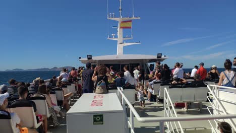 Spanish-flag-boat-full-of-seated-tourists-sailing-towards-the-Cíes-Islands-in-the-background-with-the-calm-sea-on-a-sunny-day-of-blue-sky,-shot-blocked-from-behind,-Pontevedra,-Galicia,-Spain