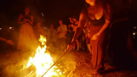 Asian-woman-managing-ceremonial-fire-with-stick-at-dance-performance-during-night,-filmed-handheld-as-wide-shot