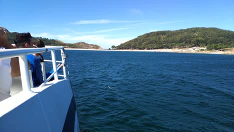 side-of-boat-with-tourists-sailing-in-front-of-the-Cíes-Islands-with-their-hills,-beach-and-forest-a-sunny-day-of-blue-sky,-blocked-shot,-Pontevedra,-Galicia,-Spain