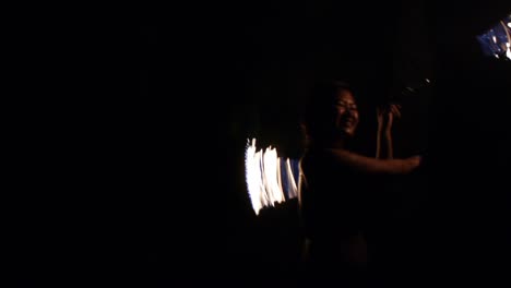 Smiling-Asian-woman-spinning-fire-ropes-at-high-speed-in-dark,-filmed-as-tight-medium-shot-from-front