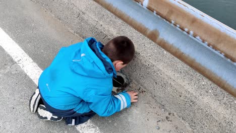 Autistic-boy-with-development-disorder-and-sensory-issues-is-stimulating-himself-by-playing-with-sand-and-gravel-and-throwing-it-down-drainhole-in-road---Handheld-static-clip-from-behind-boy