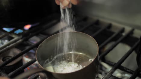 Chef-adding-salt-to-frying-onion-and-butter-in-metal-pot-on-gas-stove