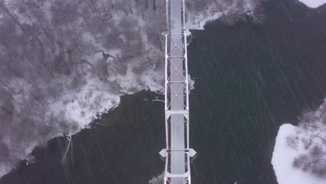 Top-down-drone-shot-of-a-bridge-crossing-a-river-in-the-middle-of-a-snow-storm