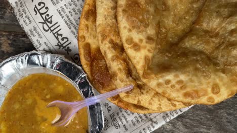 On-a-wooden-bench,-Dal-Poori-or-Puri-are-served-with-a-silver-foil-bowl-and-newspaper