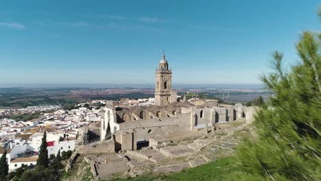 Aerial-dolly-shot-through-trees-to-the-historic-church-of-santa-maria-in-medina-sidonia-in-spain-in-the-province-of-cadiz-overlooking-the-old-town-with-white-houses-on-a-sunny-day