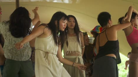 Asian-woman-wearing-white-organically-made-dress-while-dancing-ecstatically-at-music-festival-with-other-Asian-people-dancing-around,-filmed-as-medium-shot-in-slow-motion
