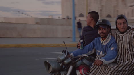 Drei-Glückliche-Arabische-Männer-Auf-Einem-Dreirädrigen-Liefermotorrad-Fahren-Im-Kreisverkehr-Vor-Der-Hassan-ii-moschee-In-Casablanca-Marokko-Und-Fahren-In-Den-Sonnenuntergang-Entlang-Der-Atlantikküste