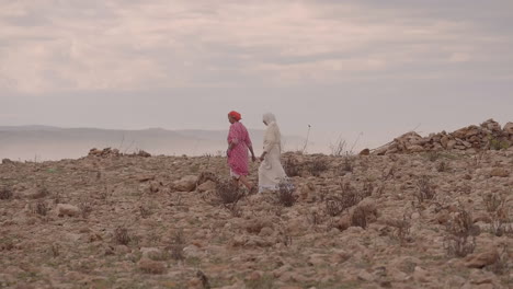 Two-women-in-traditional-Moroccan-North-African-dress-cross-a-rocky-field-on-a-cloudy,-misty-day-on-the-shore-of-the-Atlantic-Ocean