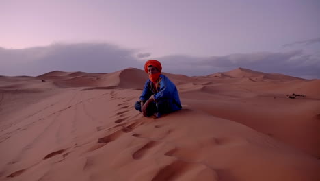 Dynamic-camera-movement-establishing-shot-proud-Berber-moroccan-man-sitting-on-sand-dunes-in-Erg-Chubby,-Merzouga,-Morocco