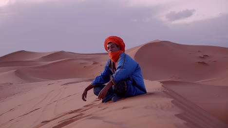 Proud-Berber-moroccan-man-looking-at-camera-and-into-the-distance,-sitting-on-sand-dunes-in-Erg-Chubby,-Merzouga,-Morocco