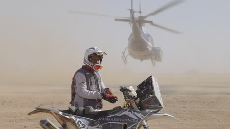 Radfahrer,-Der-Während-Der-Dakar-rallye-Handschuhe-In-Staubiger-Umgebung-Anzieht,-Während-Der-Hubschrauber-Im-Hintergrund-Abhebt