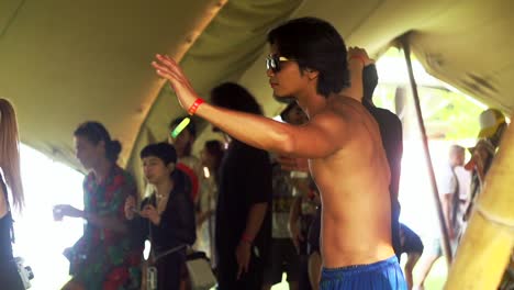 Shirtless-Asian-man-wearing-black-sunglasses-with-arms-swinging-around-in-dancing-motion-during-ecstatic-dance-movement,-filmed-as-medium-tight-shot-in-slow-motion