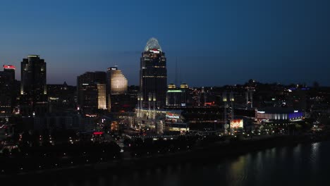 Aerial-view-over-the-Ohio-river,-towards-the-illuminated-Great-American-Ball-Park,-dusk-in-Cincinnati,-USA---rising,-drone-shot