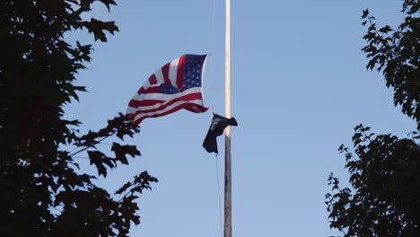 Bandera-Estadounidense-A-Media-Asta-Ondeando-Junto-Al-Emblema-Oficial-De-La-Liga-Nacional-De-Familias-Pow-Mia-Flag-En-Central-Park,-Estableciendo-Una-Vista-De-ángulo-Bajo-A-Través-De-Ramas-De-árboles,-Día-Conmemorativo