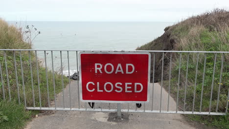 Waves-roll-in-on-the-North-Sea-behind-a-red-road-closed-sign-and-barrier-warns-of-where-coastal-erosion-has-destroyed-a-road-on-the-Norfolk-coastline