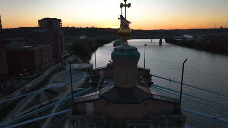 Aerial-view-over-the-flag-on-the-Roebling-Bridge,-sunset-in-Covington,-Kentucky,-USA---reverse,-drone-shot