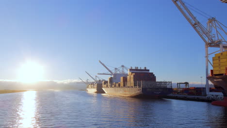 Low-angle-aerial-view-passing-Oakland-port-container-ships-and-industrial-gantry-crane-on-shimmering-waterfront
