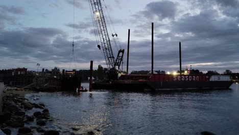 Team-Of-People-Working-At-Sunset,-Dismantling-Massive-Barge-Washed-Ashore-By-Storm-On-The-Seawall-Of-English-Bay-In-Vancouver,-Canada