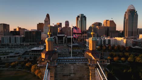 Aerial-view-around-the-US-Flag-waving-on-the-Roebling-bridge-with-sunlit-skyscrapers-in-the-background,-in-Cincinnati,-USA