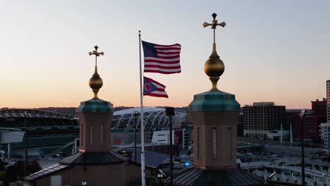 Aerial-view-around-the-US-and-Ohio-flag-on-Roebling-bridge-with-sunset-Cincinnati-skyline-background---circling,-slow-motion-drone-shot