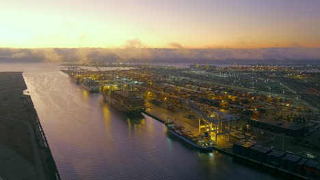 Port-of-Oakland-at-twilight-with-lights-glowing-as-containers-are-loaded-and-unloaded-from-cargo-ships---pull-back-aerial-reveal