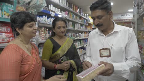 Ein-Verbraucherbeauftragter-Weist-Zwei-Indische-Frauen-Mittleren-Alters-Beim-Einkauf-Von-Einzelhandelsartikeln-Auf-Irreführende-Verpackungspraktiken-Hin