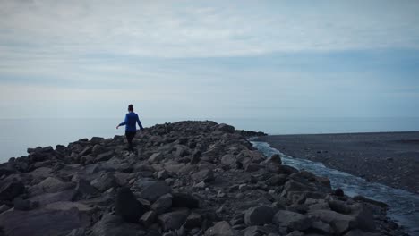 Tiro-De-Seguimiento-De-Niña-Mujer-Caminando-Con-Cuidado-Sobre-Rocas-Volcánicas-Negras,-Playa-Negra,-Islandia