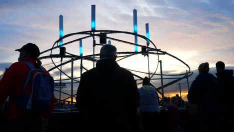 Silhouette-audience-interact-with-illuminated-spiral-light-looper-neuron-artwork,-Liverpool-pier-head-river-of-light-event-at-sunset