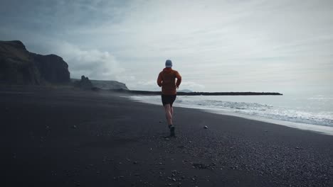 Low-Angle-Tracking-Shot-Of-Fit-Man-Wearing-Red-Shirt-Running-On-Reynisfjara-Black-Beach,-Iceland