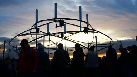 Silhouette-of-public-interact-with-illuminated-spiral-light-looper-neuron-artwork,-Liverpool-pier-head-river-of-light-event-at-sunset
