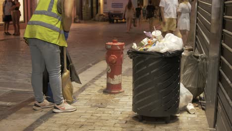 Female-street-sweeper-sweeping-up-waste-from-overflowing-rubbish-bin