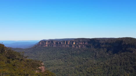 Dramatic-rising-aerial-drone-flyover-of-rock-outcrop-covered-in-trees-and-bushes-in-the-Blue-Mountains-clear-blue-sky-in-New-South-Wales
