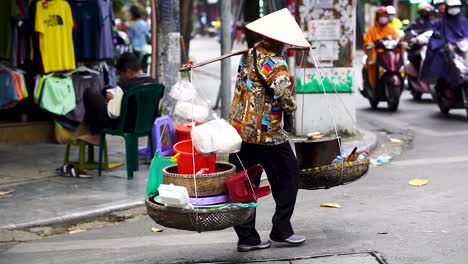 Traditionelle-Vietnamesin,-Die-Waren-Auf-Körben-Auf-Der-Schulterkreuzungsstraße-In-Hanoi-Trägt
