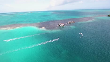 Zwei-Männer-Kitesurfer-überqueren-Das-Meerwasser-Fliegend,-Drohnenschuss-Palafito-Los-Roques