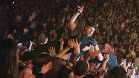 Spectators-Crowd-Cheering-Arms-Raised-Carrying-Man-During-Music-Concert-Show-in-Arena,-Security-Guy-Intervention,-Stands-and-Audience-Around-with-Spotlights
