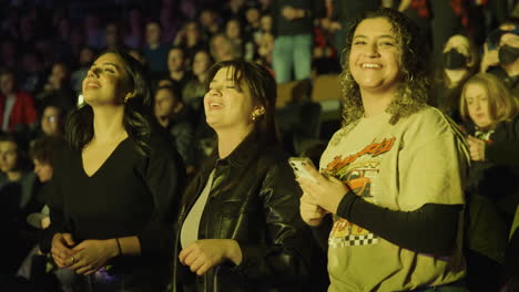 Joyful-Women,-Three-Young-Ladies-Spectators-in-Arena-Stands-During-Music-Concert-Show,-Smiling-Singing-and-Nod-Heads,-Crowd-Audience-Around